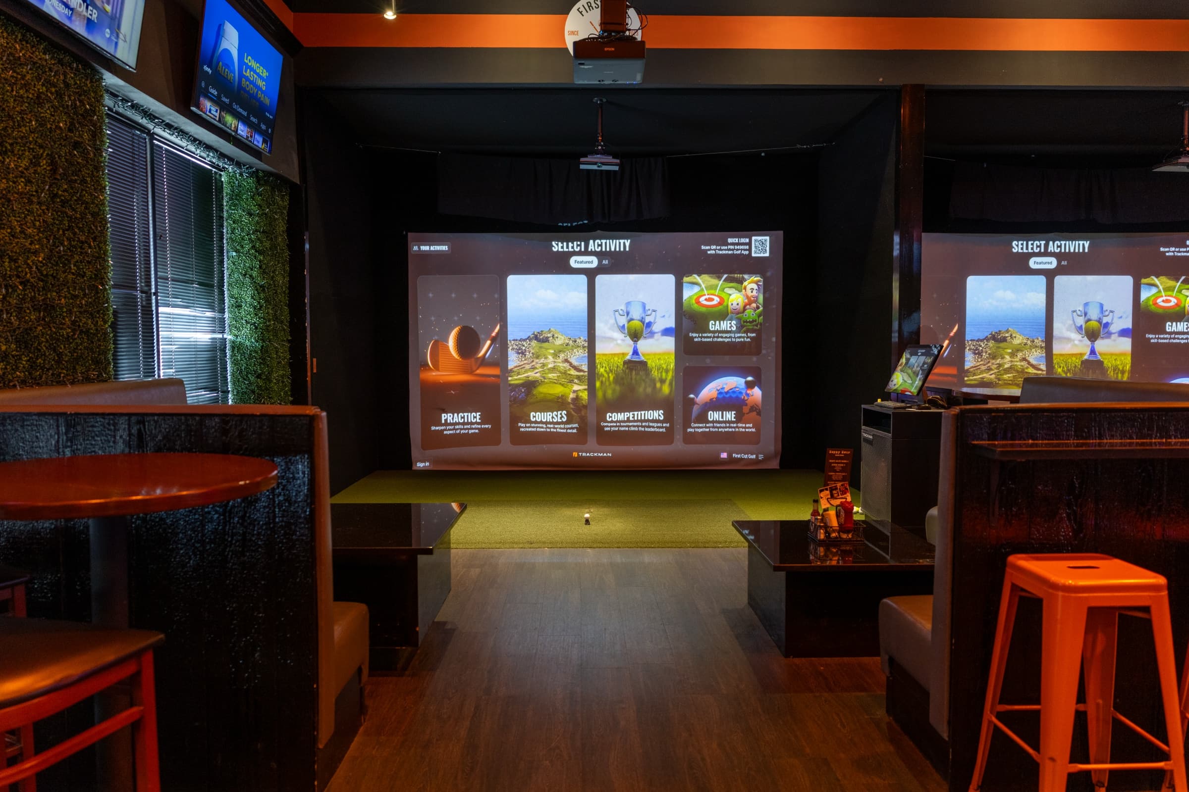 A simulator bay between two booth tables at First Cut Golf, with the TrackMan IO screen showing the activity menu and orange brand stools flanking the walkway.